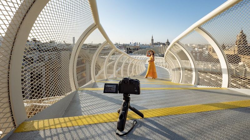 אישה בשמלת קיץ צהובה מצטלמת על גשר הולכי רגל מתעקל, עם Canon EOS R10 על חצובה קטנה הממוקמת במיוחד כדי לצלם אותה.