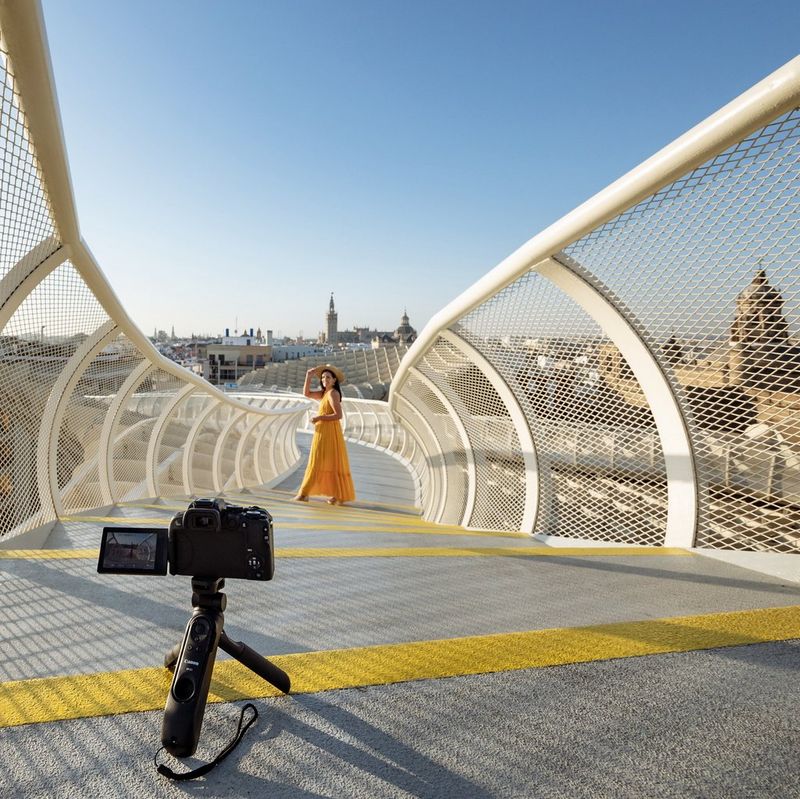 A Canon EOS R10 on a tripod in the foreground points towards a woman in the middle distance on a curving walkway, with a city in the background.