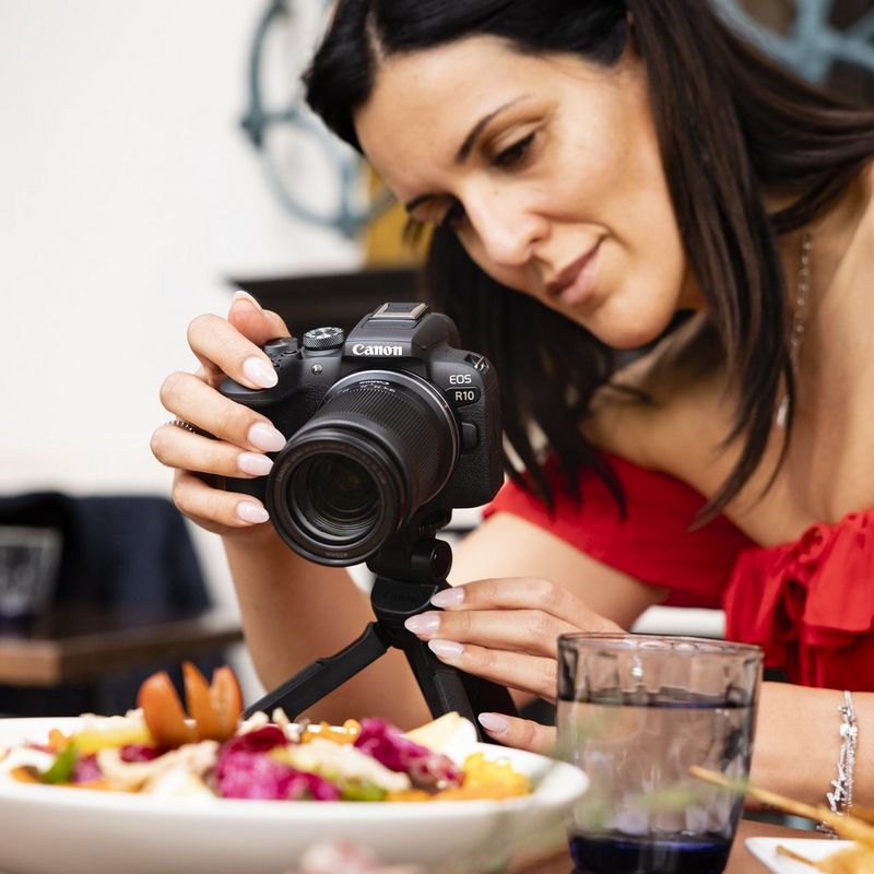 A woman photographs a plate of food on a table using a Canon EOS R10 camera with an RF-S 18-150mm F3.5-6.3 IS STM lens.