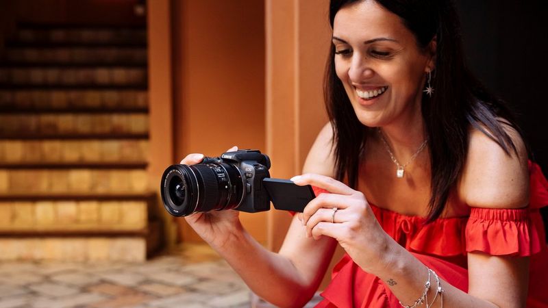 A woman wearing a red strapless dress crouches down to take a picture with a Canon EOS R10.