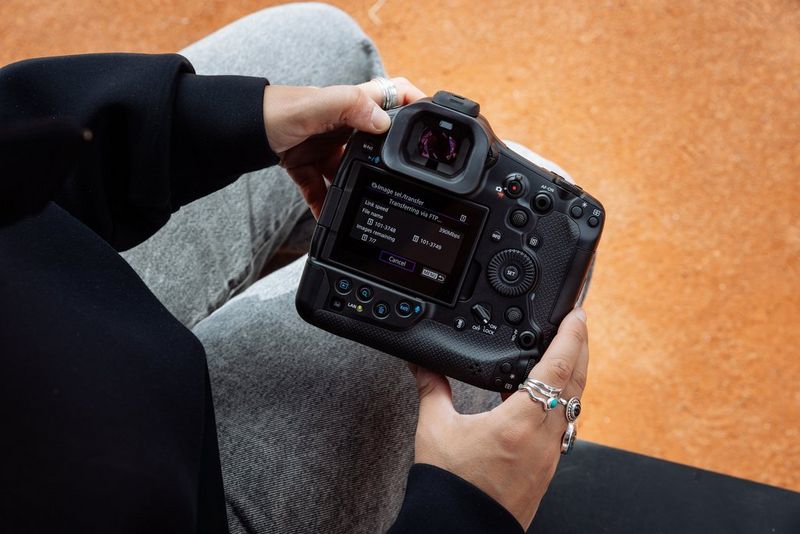 A user at the side of a clay tennis court holds a Canon EOS R1 camera. The screen shows an FTP file transfer taking place.