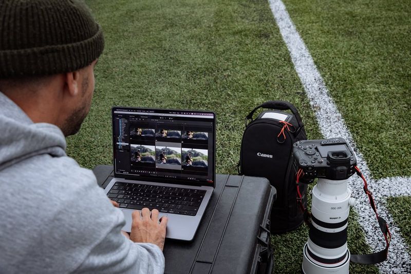  A man reviews a set of images on a laptop resting on a hard camera case on a football pitch, alongside a Canon EOS R1 with a telephoto lens. 