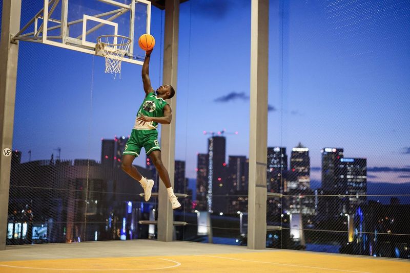 Een basketbalspeler midden in zijn sprong, reikend naar de ring tegen de achtergrond van een donkere stadsskyline. Gefotografeerd met een Canon EOS R1.