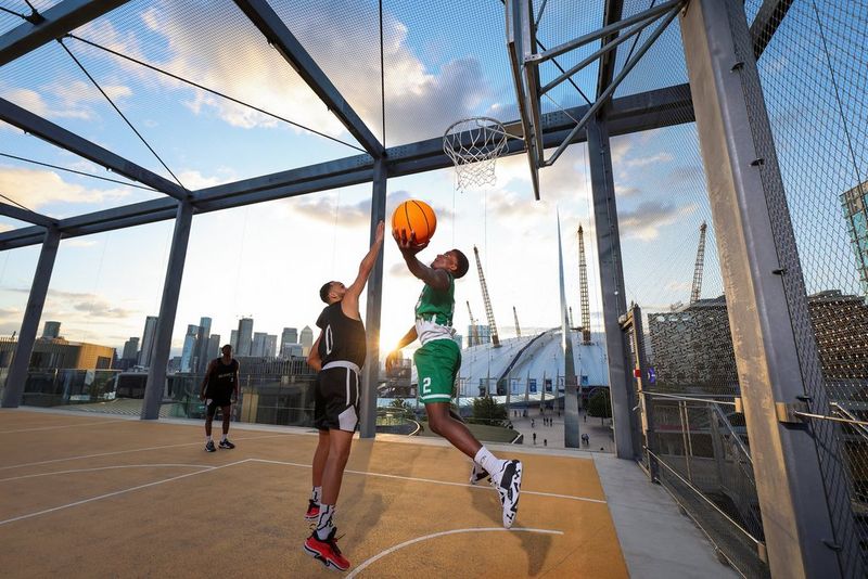 Op een foto die is gemaakt met een Canon EOS R1 zijn twee jonge mannen aan het basketballen, de een in het zwart en de ander in het groen, terwijl ze proberen te schieten. Ze spelen op een dakterras met de ondergaande zon en de skyline van de stad erachter.