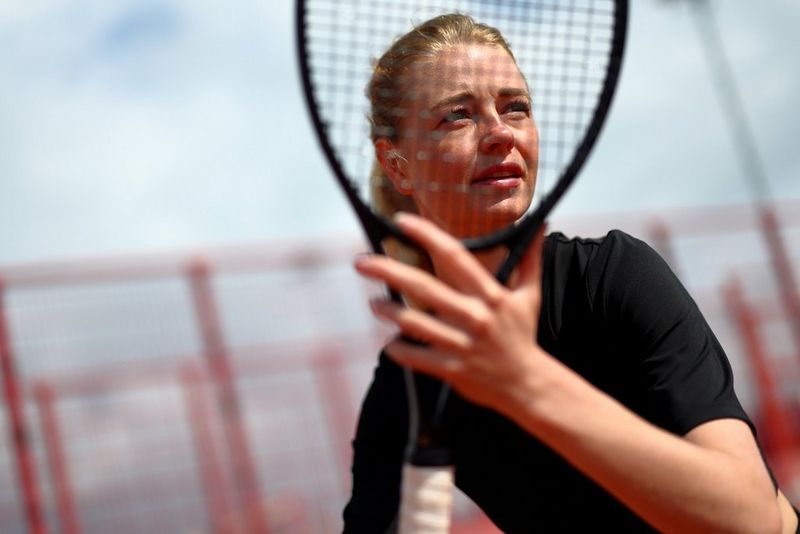 A portrait of a tennis player with a focused expression, photographed through her racket, with the fence around the court blurred in the background under a partly cloudy sky.