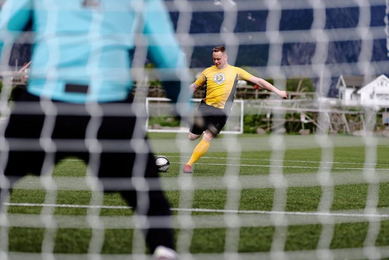 Фото через сетку ворот, созданное на Canon EOS R1; на снимке футболист в полном фокусе готовится ударить в сторону ворот, а на переднем плане можно увидеть спину вратаря.