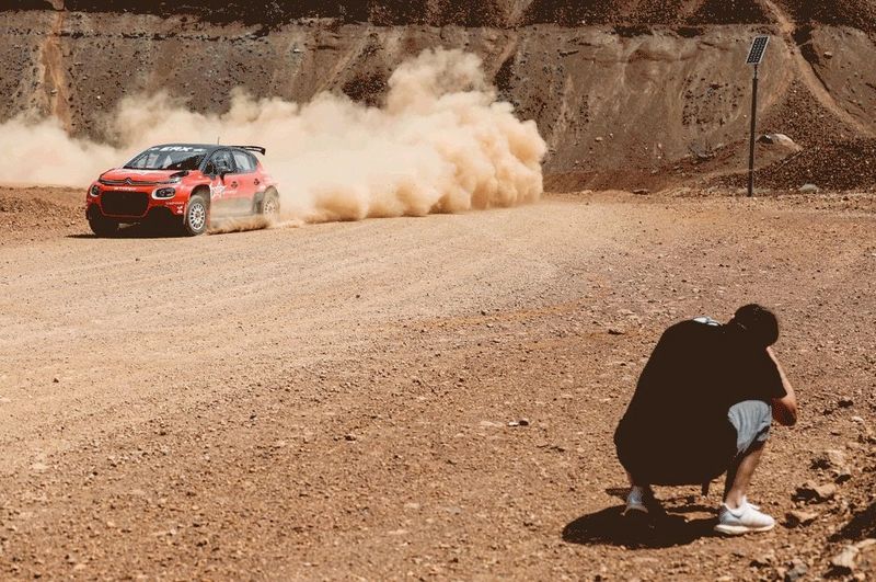 A man crouches down to photograph a rally car as it rounds a corner shrouded in a cloud of dust.