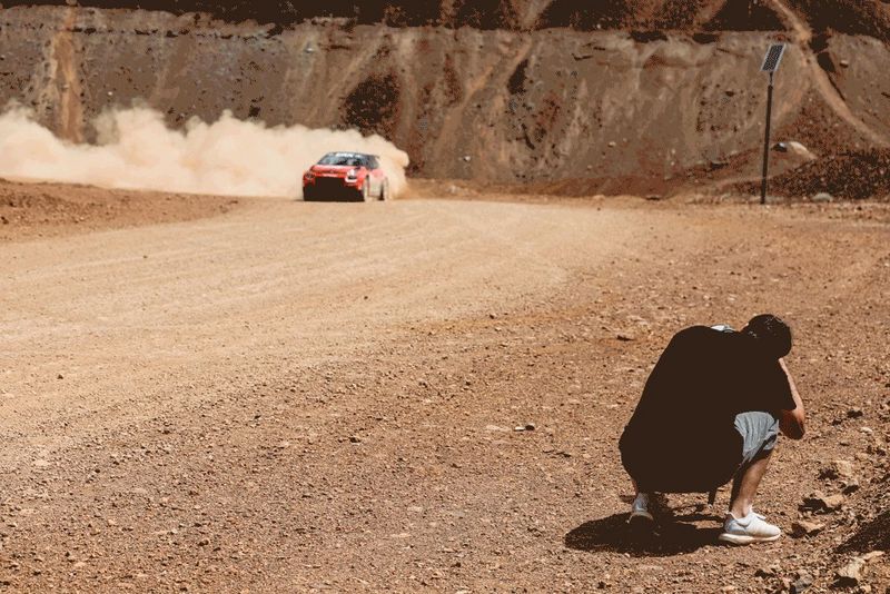 Мужчина присел, чтобы запечатлеть раллийный болид, выезжающий из-за поворота в облаке пыли.