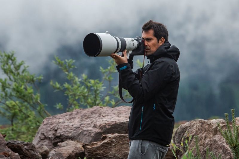 Un uomo con una giacca nera, immerso in un panorama di nuvole basse sopra le rocce, scatta a mano libera utilizzando una fotocamera Canon EOS R3 e un super teleobiettivo.