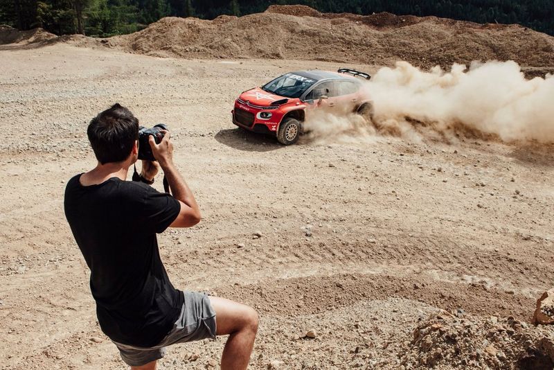 A man takes pictures of a red rally car as it swerves around a corner, kicking up dust from its tyres.