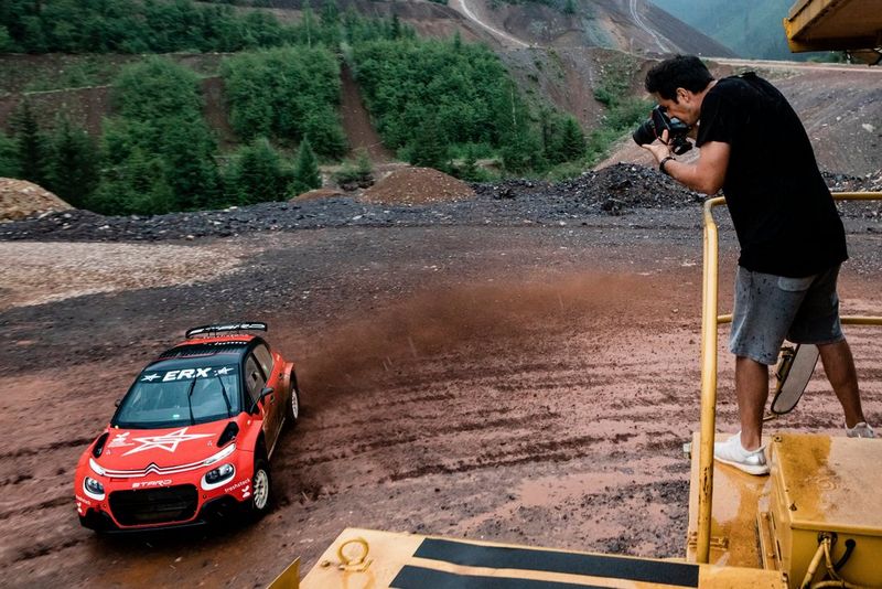 Un homme debout sur un portique surélevé avec un appareil photo à ses yeux regarde en direction d'une voiture de rallye rouge située au sol.