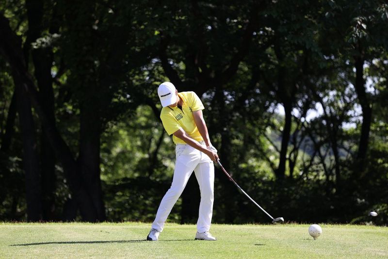 A golfer in white trousers and yellow shirt captured just after striking the ball. The shaft of the club looks curved.