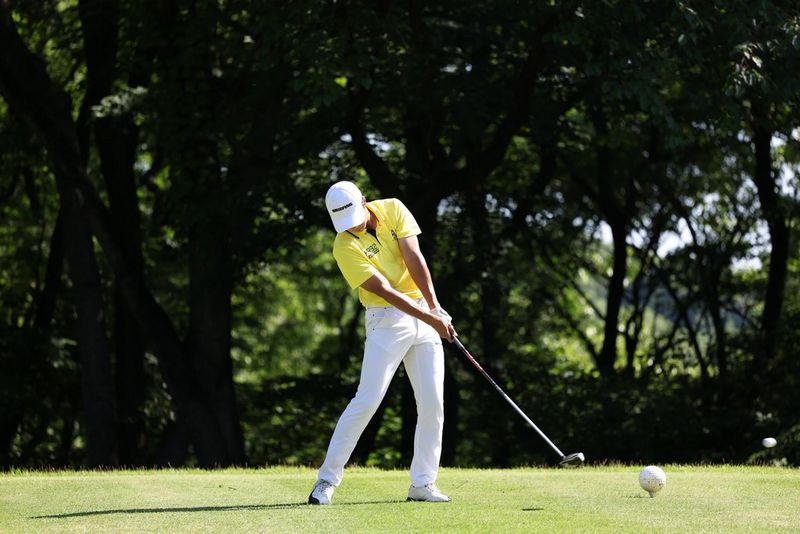 The same golfer in white trousers and yellow shirt captured at the same moment, just after striking the ball. The shaft of the club looks straight.