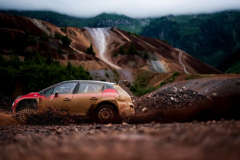 Une voiture de rallye rouge sale filant à toute allure à travers l'image : elle soulève un nuage de poussière derrière elle, l'arrière-plan montagneux est flouté.