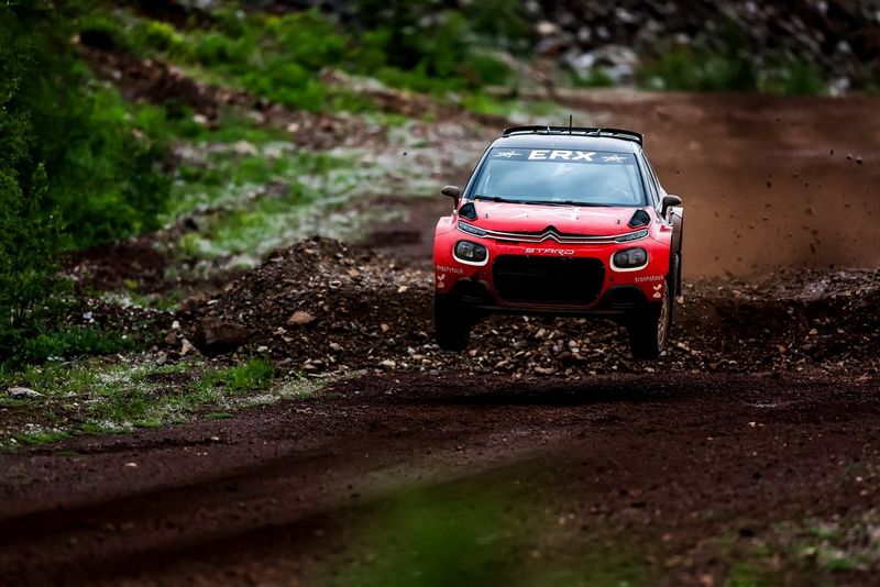 Un'auto da rally rossa sfreccia a tutta velocità, con le quattro ruote sollevate da terra, dopo aver superato un dosso, sulla pista sterrata.