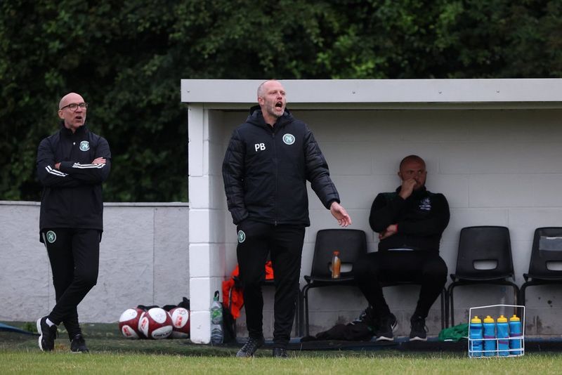 Een man in een zwart jack en zwarte broek schreeuwt terwijl hij naar een voetbalwedstrijd kijkt. Er staat nog een man naast hem, met zijn armen over elkaar, terwijl rechts op de foto een derde man in een dug-out zit met een mand drinkflessen bij zijn voeten.