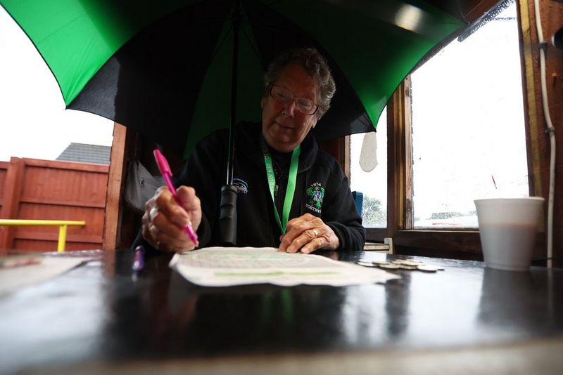 Een vrouw met een open groen-zwarte paraplu zit in een houten hokje en schrijft op een vel papier.