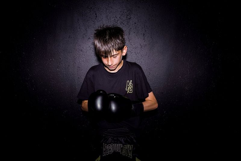 Een portret van een jonge kickbokser tegen een donkere achtergrond, verlicht met een flitser. De jongen kijkt omlaag naar zijn handen in kickbokshandschoenen die hij stevig tegen elkaar duwt.