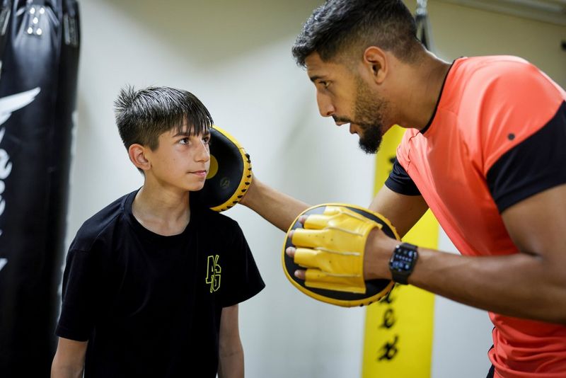Een mannelijke bokser met handpads buigt zich voorover om te praten met een jongen in een zwart T-shirt toe die tegen hem opkijkt.