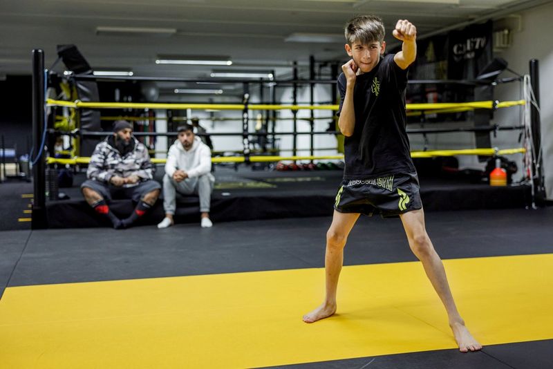In a photo taken on a Canon EOS R3 by Nikolai Linares, a young boxer practises punching as two men look on, sitting against the yellow ropes at the edge of the ring, out of focus in the background.