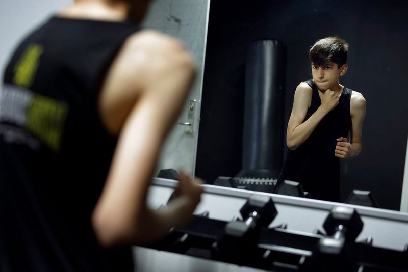 Een jongen spant zijn biceps aan voor een rek met gewichten. In de spiegel achter de gewichten is zijn weerspiegeling te zien.