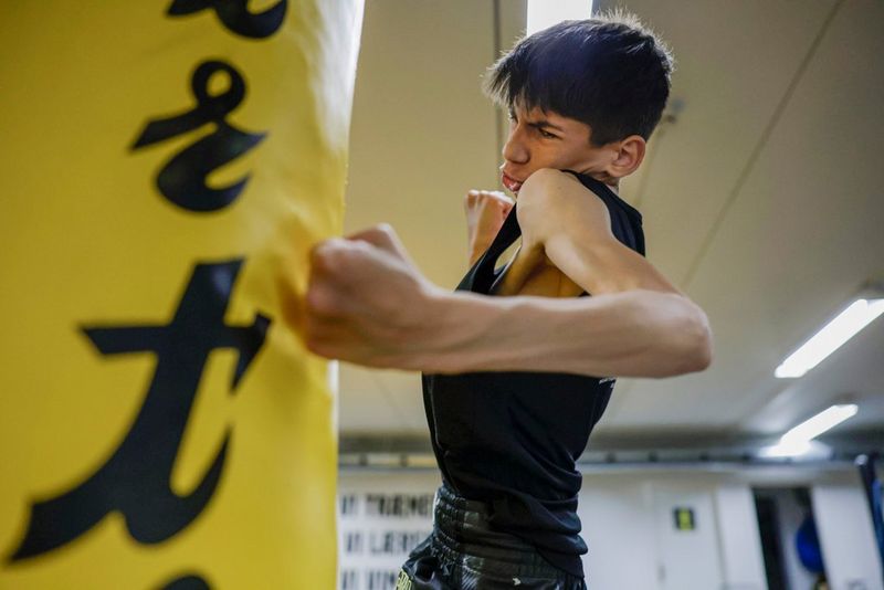 Een jongen is gefotografeerd tijdens een stoot tegen een gele bokszak, waarbij hij zijn spieren aanspant.