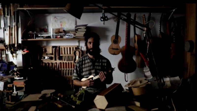 A bearded man stands in a workshop holding a small wooden ukelele, with others of different sizes hanging on the wall behind him.