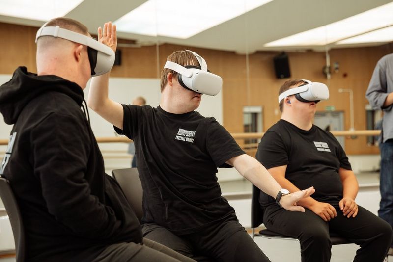 Three young men sit in chairs wearing VR headsets. The middle of the men is stretching one arm forward and the other up, in imitation of the ballet he is watching.