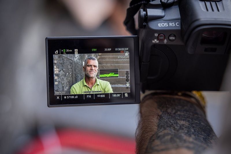 Una videocamera ibrida Canon EOS R5 C con lo schermo posteriore che mostra un uomo con una t-shirt verde, e varie impostazioni del dispositivo selezionate.