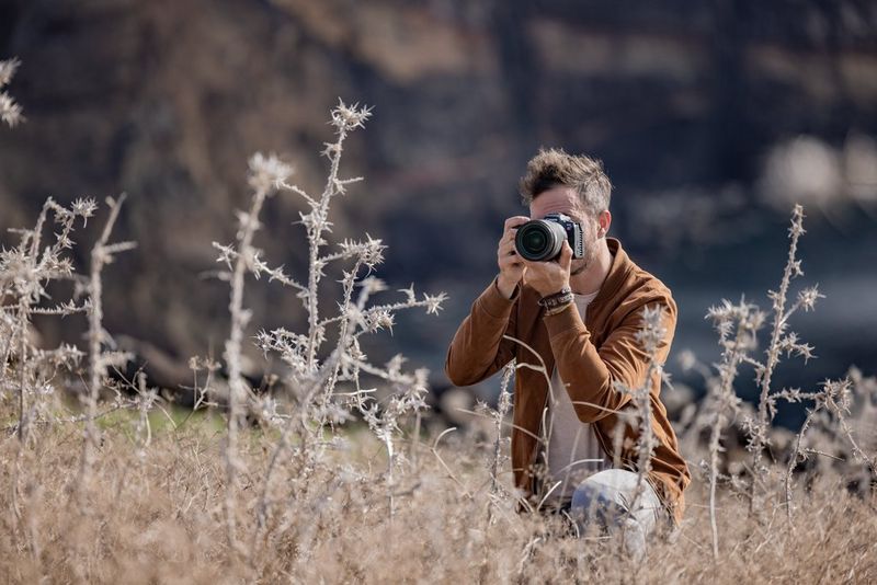 A photographer kneels among dry plants while taking a photograph with a mirrorless camera.