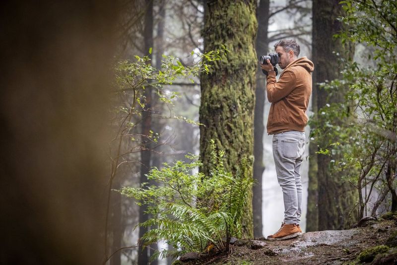 A man in a tan jacket and grey jeans filming in a woodland setting with a Canon EOS R5 C camera.
