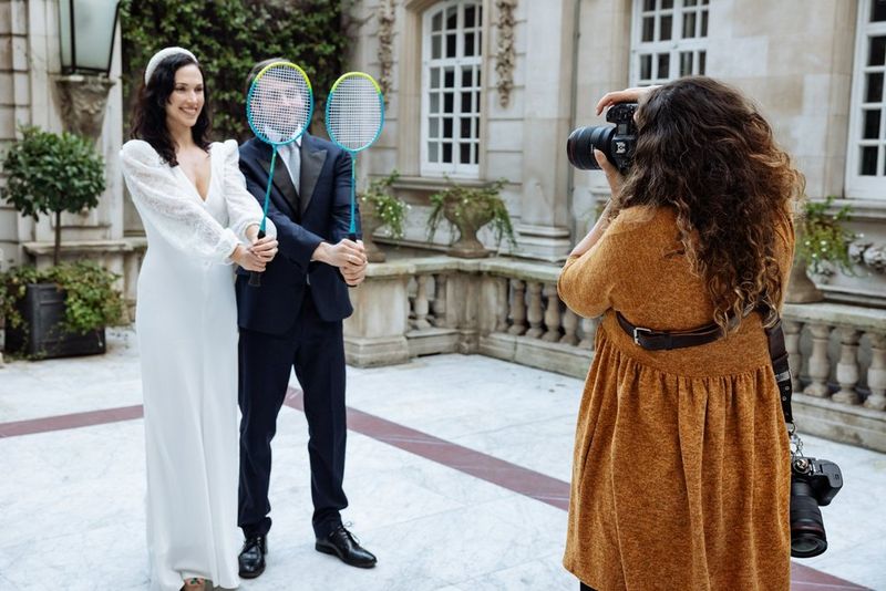 Una sposa e uno sposo tengono delle racchette da badminton davanti al viso mentre vengono fotografati da Alison Bounce.
