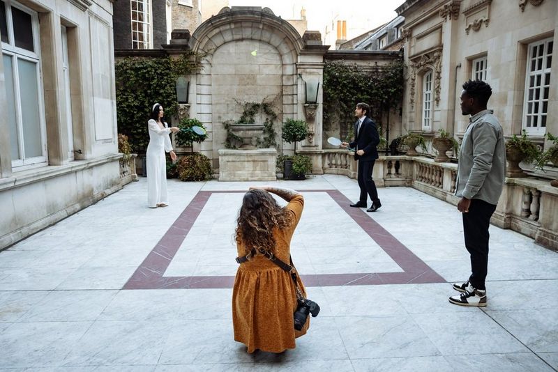Photographer Alison Bounce crouches down to photograph a bride and groom playing badminton in a courtyard.