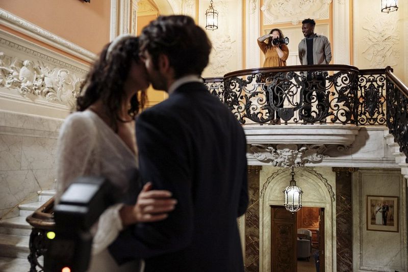La fotografa Alison Bounce è in piedi su una scalinata con un abito arancione e tiene in mano la fotocamera per fotografare una coppia che si bacia in primo piano.