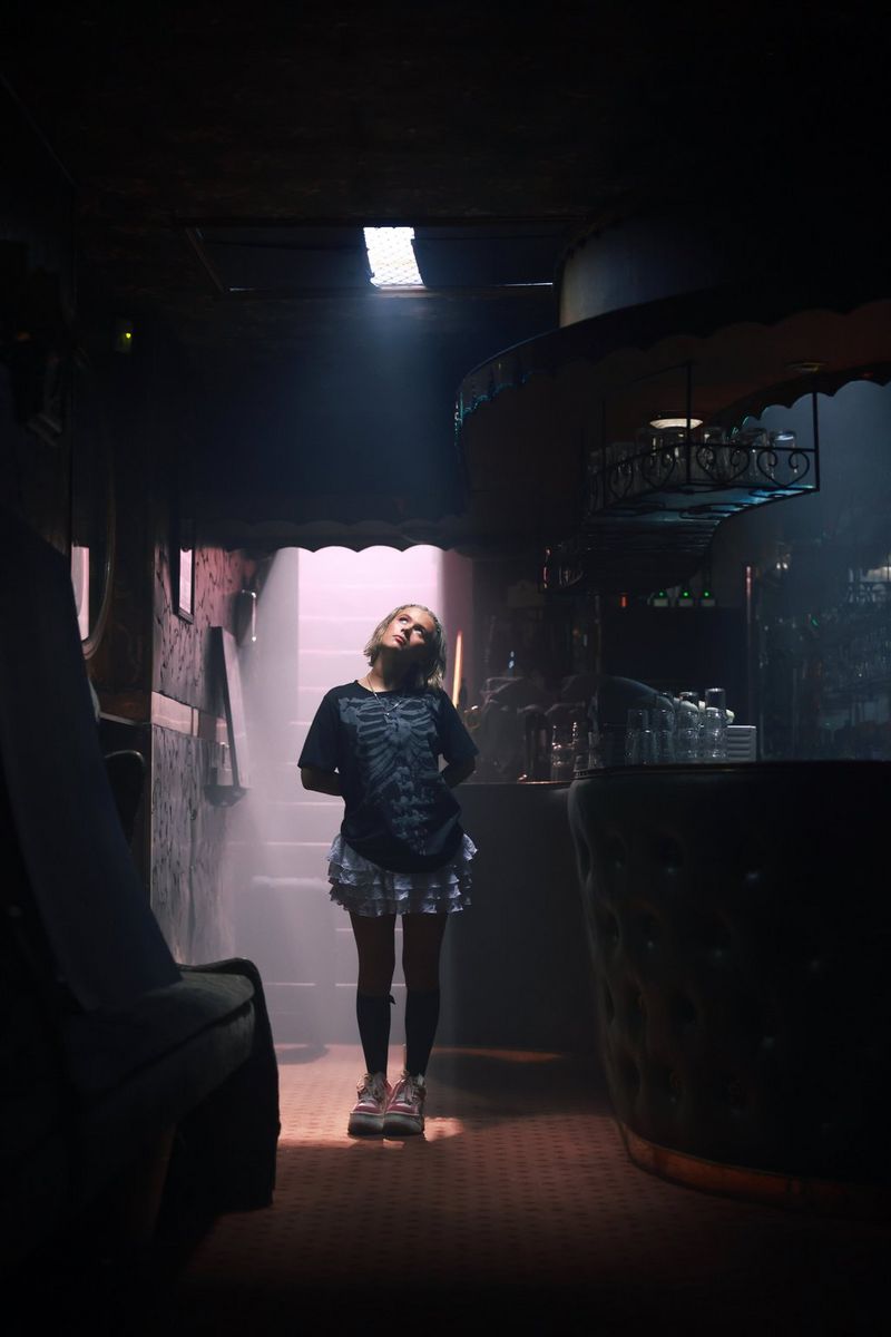 In a low-light photo taken on a Canon EOS R5 Mark II, a person stands in a dimly lit bar illuminated by the light from a small skylight and a staircase behind her.
