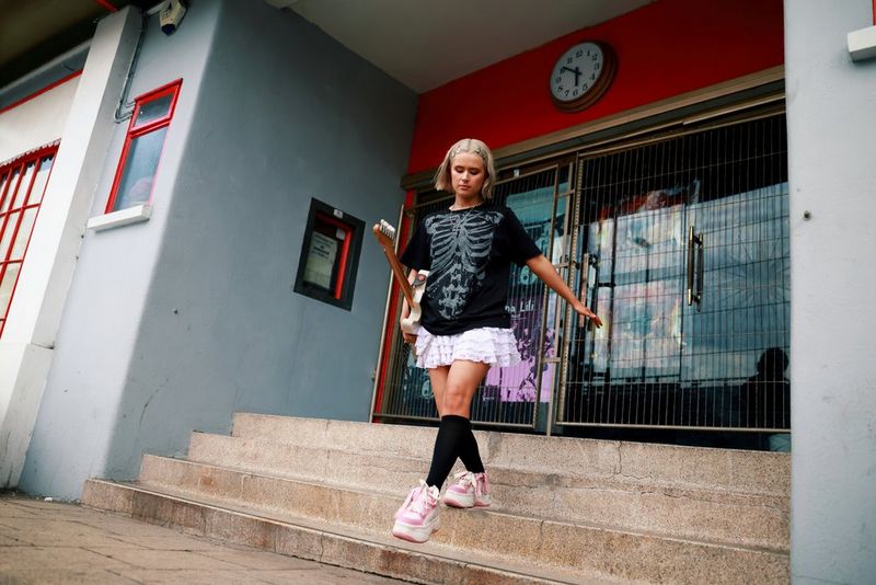 In a photo taken with a Canon RF 28-70mm F2L USM lens, a woman wearing a dark top and white skirt walks down the steps of a building, carrying a guitar in one hand.