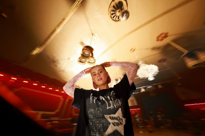 Девушка стоит в концертном зале и смотрит в объектив, обхватив руками голову; фон изображения немного искажен и размыт. Снято на Canon EOS R5 Mark II.