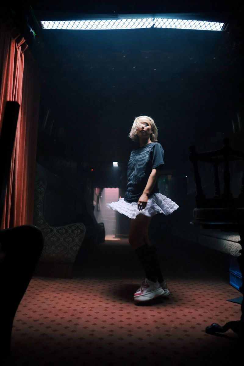 Dekle v majici in krilu se v slabo osvetljenem prostoru obrne proti kameri.