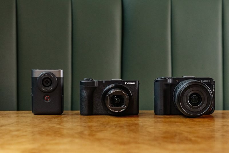 Камеры Canon PowerShot V10, PowerShot V1 и EOS R50 V стоят на деревянном столе, за которым находится зеленый кожаный диван.