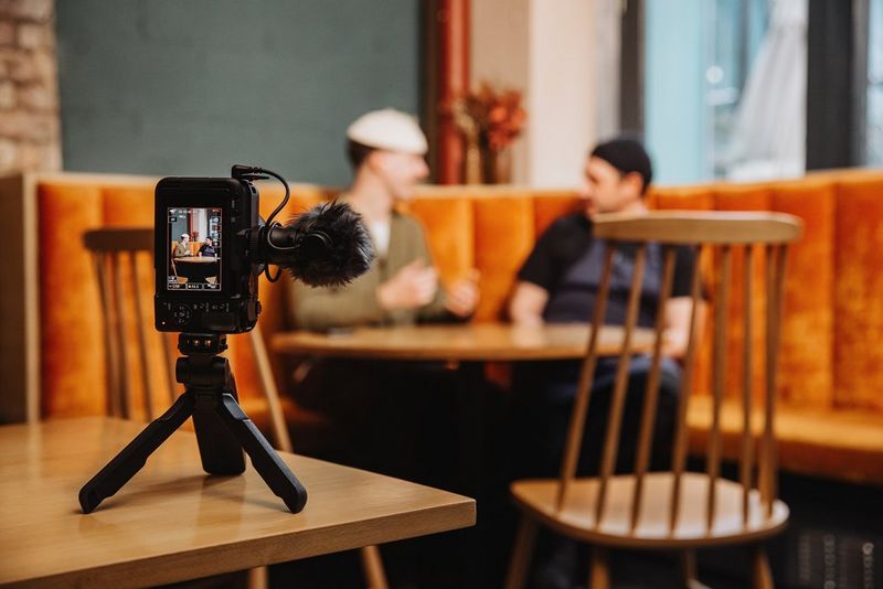 Fotoaparát Canon EOS R50 V s připevněným externím mikrofonem upevněný na stativu na stole a natáčí dvě osoby sedící na rohovém sedátku na pozadí.