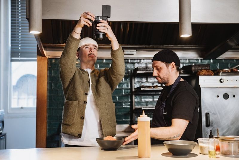 In a kitchen, a vlogger films a bowl of food from above while the chef sits to one side, one hand touching the bowl.