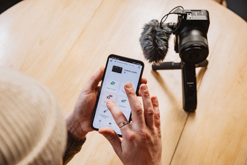 A Canon EOS R50 V camera with a microphone attached, being prepared for a livestream by a man using a smartphone.