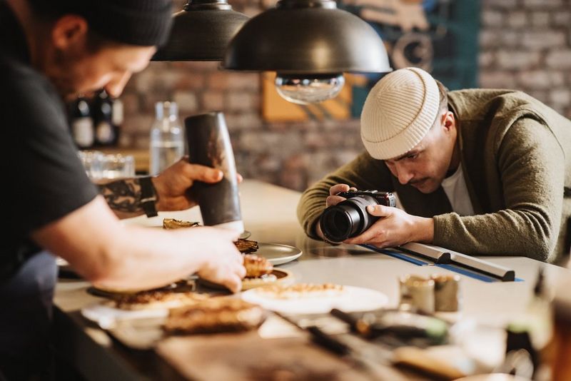 A user holds a Canon EOS R50 V at worktop height, filming a chef preparing a plate of food.