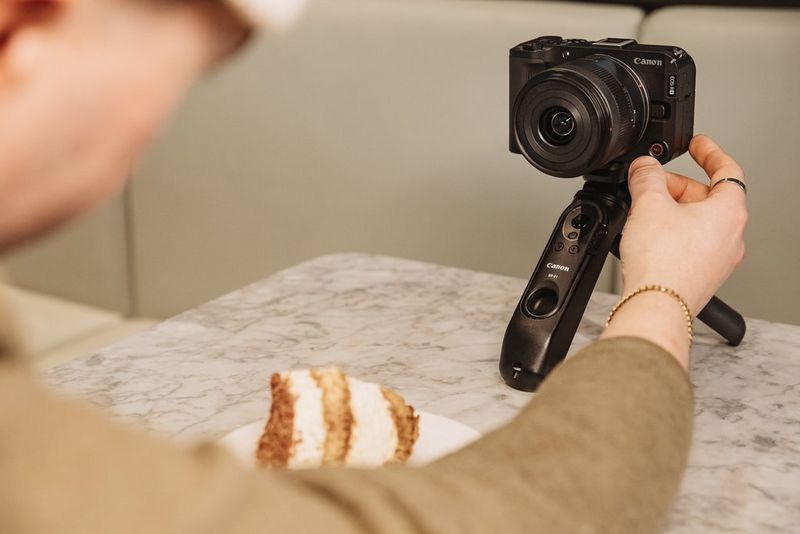 A user reaches for the record button on the front of a Canon EOS R50 V facing him and a slice of cake in front of him.
