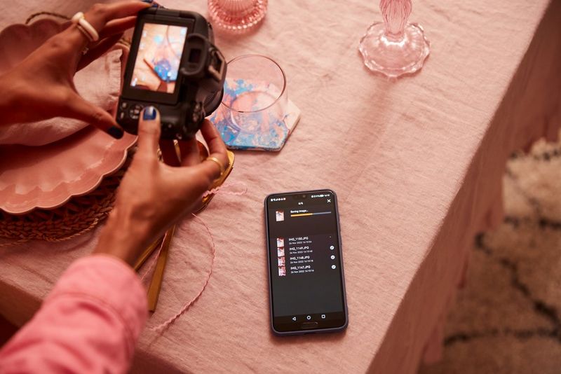 Delle mani reggono Canon EOS R50 durante il trasferimento di file dalla fotocamera a un telefono posato sul tavolo accanto.