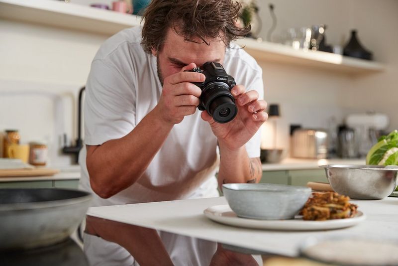Food influencer Martyn Odell leans over a kitchen counter with the Canon EOS R50, adjusting the lens to film a plate of food.
