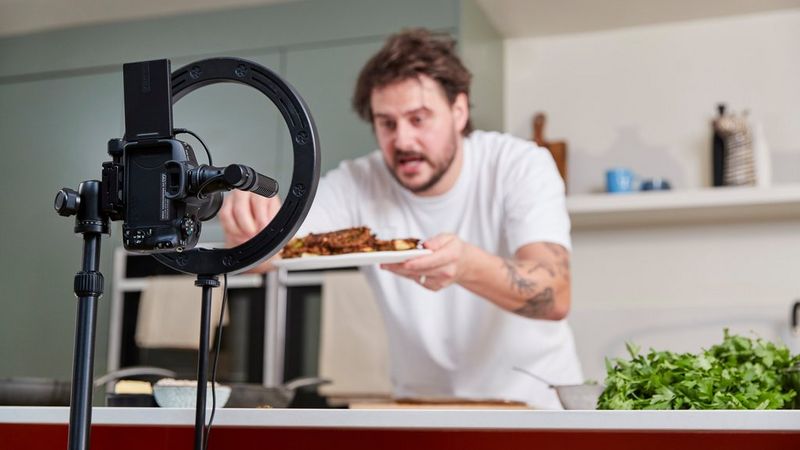 A content creator, Martyn Odell, carefully plates food in a kitchen while being filmed. The Canon EOS R50 is set up on a tripod with a ring light in the foreground, capturing the cooking process.
