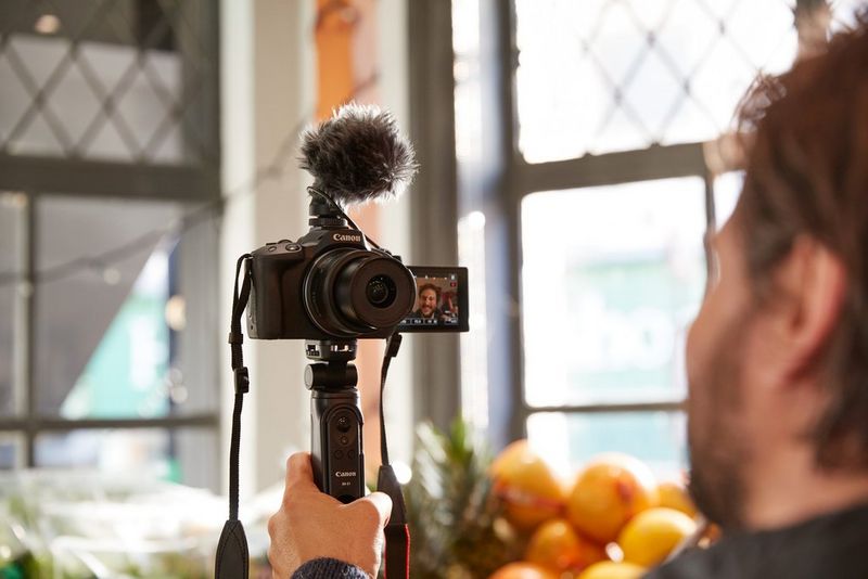 Una mano stringe lo stabilizzatore per treppiede HG-100TBR di Canon collegato a Canon EOS R50 con un microfono stereo Canon DM-E100.