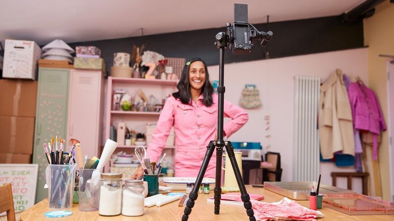 Canon EOS R50 su un treppiede posizionato sopra un tavolo per riprendere una donna in tuta rosa circondata da strumenti per il fai da te.