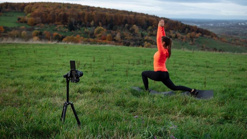 A woman in a bright orange top performs a yoga pose on a mat in a lush green field with a scenic view of autumnal trees and distant hills. A Canon EOS R50 camera is positioned on a tripod in the foreground, capturing the scene.